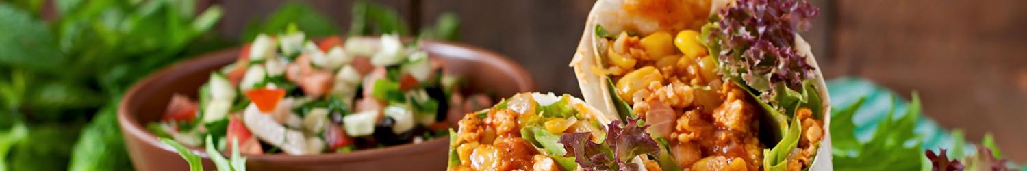 Nahaufnahme eines Wraps gefüllt mit Salat, Mais und gewürfeltem Gemüse, vor einer Schüssel Salat mit Tomaten und Gurken. Grüne Kräuter sind auf einer Holzoberfläche verstreut.