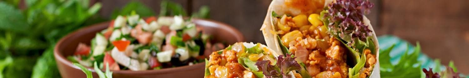 Eine Nahaufnahme eines mit Mais, Salat und gekochtem Fleisch gefüllten Burritos, vor einer Schüssel Salat mit Gurke, Tomate und Grünzeug platziert.