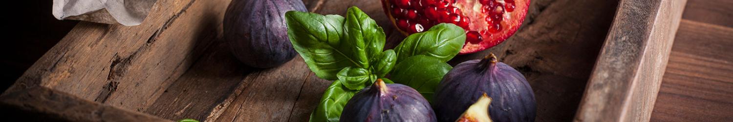 A wooden tray with figs, a cut pomegranate, and basil leaves on a rustic surface.