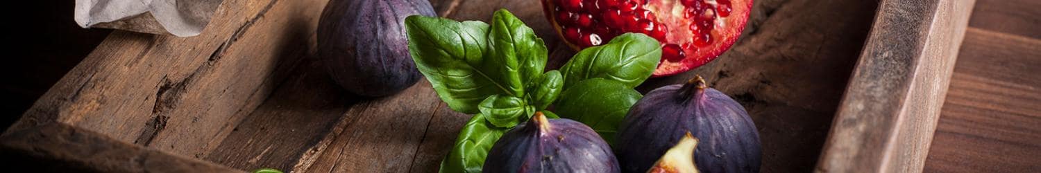 A wooden tray with figs, a cut pomegranate, and basil leaves on a rustic surface.