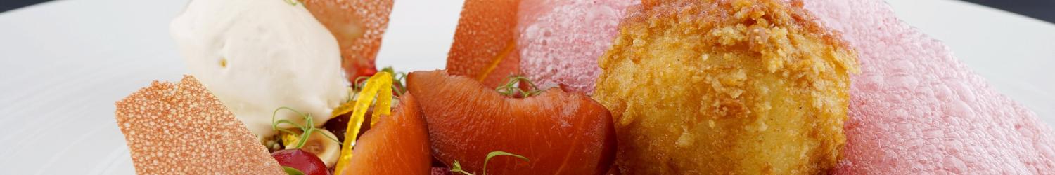Ein Gourmet-Dessert mit einer frittierten Kugel, rosa Schaum, einer Kugel Eis, roten Fruchtscheiben, Haselnüssen und dünnen knusprigen Stücken auf einem weißen Teller.