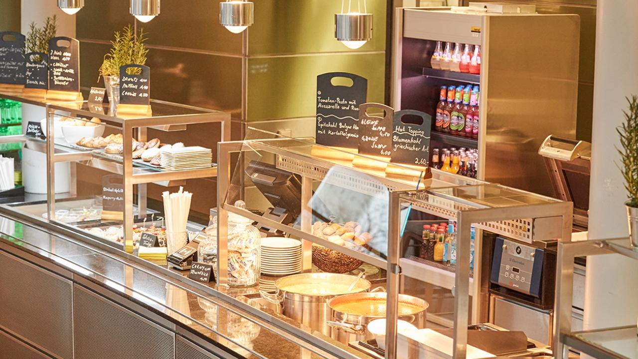 Cafeteria counter with a variety of packaged snacks, bottled drinks, and a hot food section. Several chalkboard signs display menu options and prices.