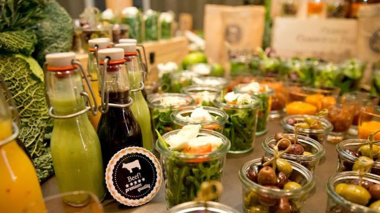 Verschiedene Gläser mit Salaten, frischen Säften und eine Flasche Balsamico-Dressing, arrangiert auf einem Tisch mit Blattgemüse im Hintergrund.
