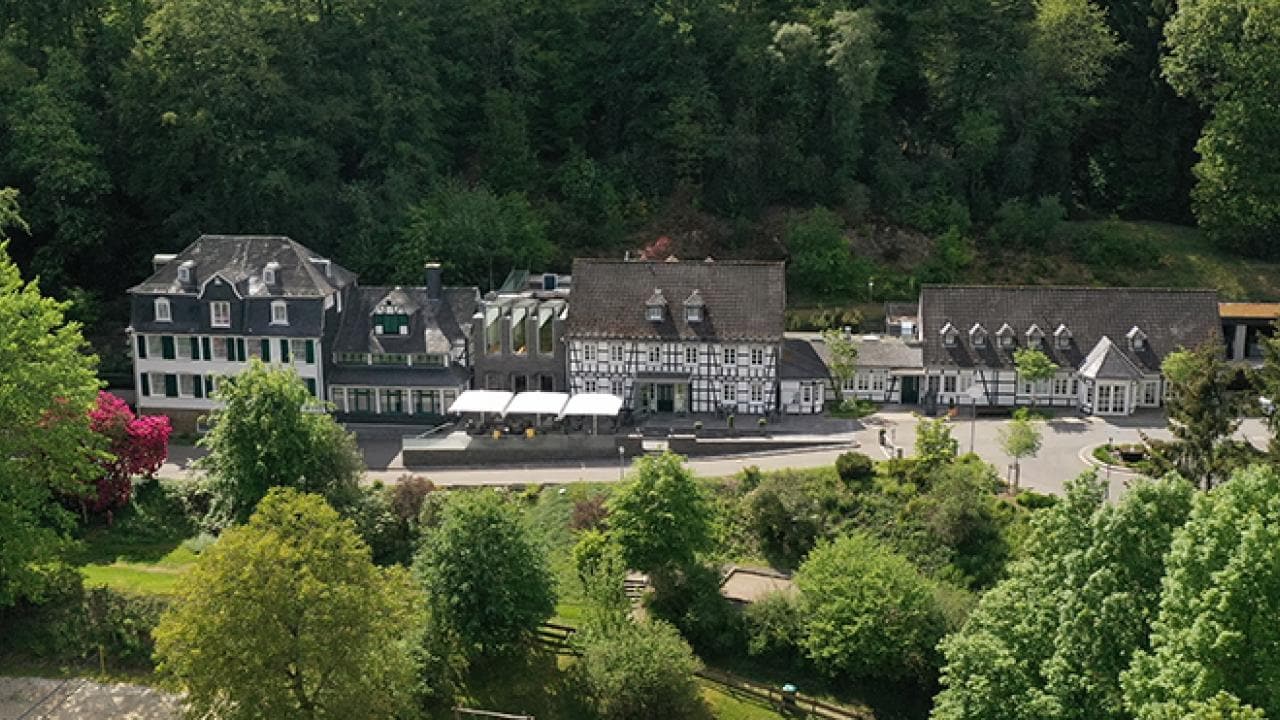 Aerial view of a country estate surrounded by lush greenery, featuring several connected buildings with a mix of white and dark roofs.