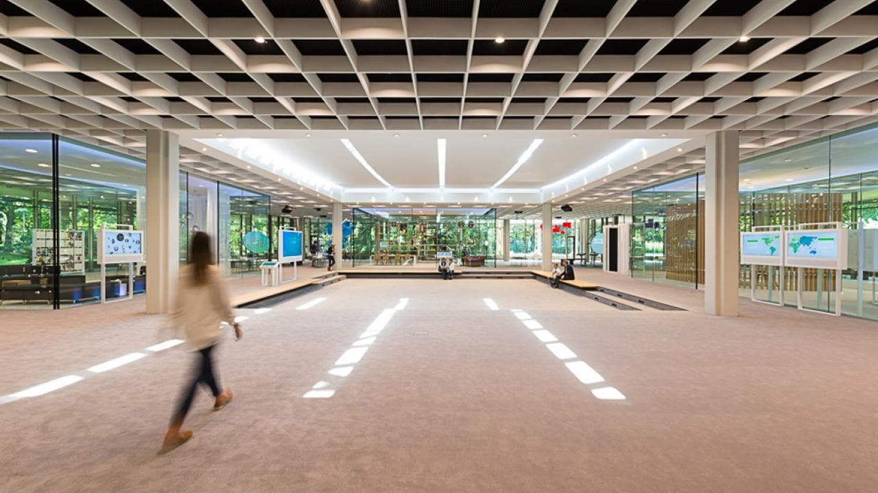 Spacious modern office lobby with geometric ceiling, glass walls, and digital displays. A person walks across the carpeted floor.