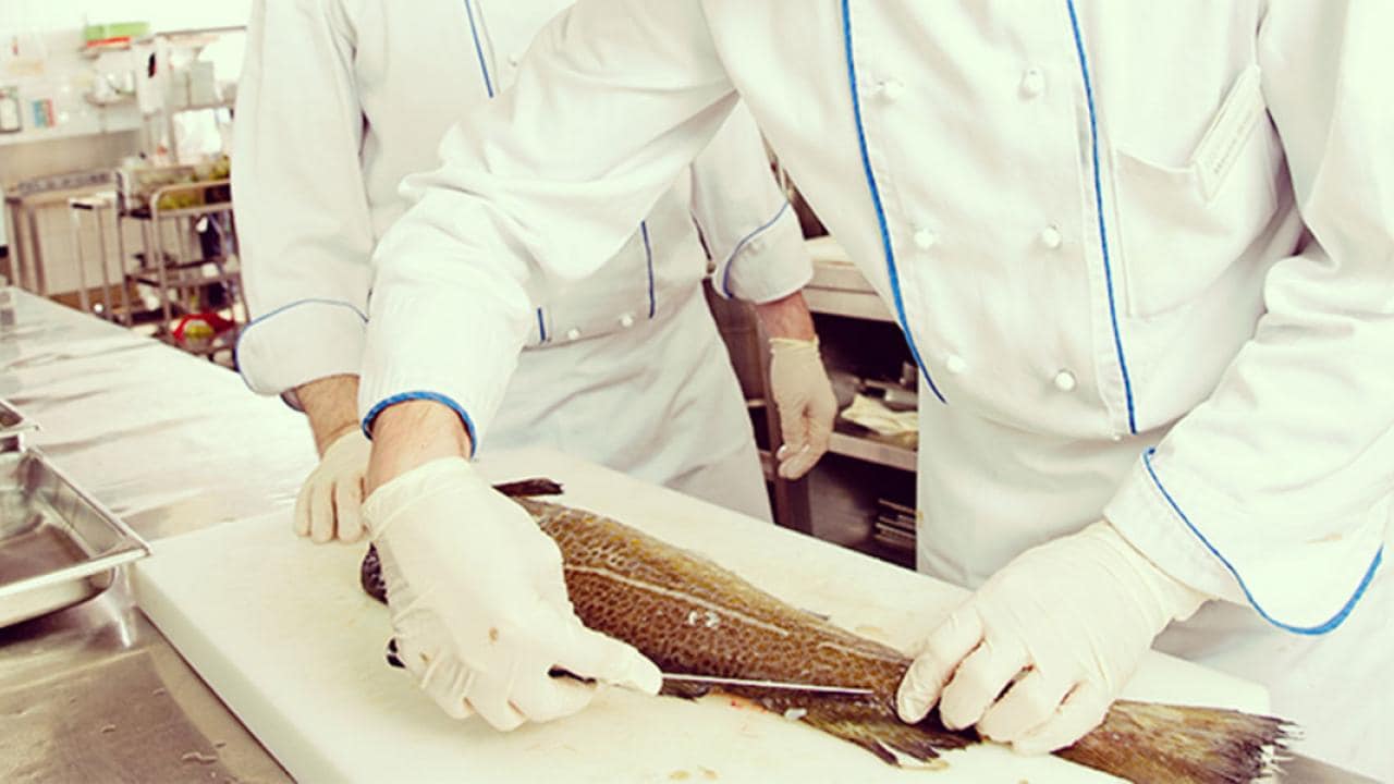 Chefs in a kitchen preparing raw fish on a cutting board, wearing white uniforms and gloves.