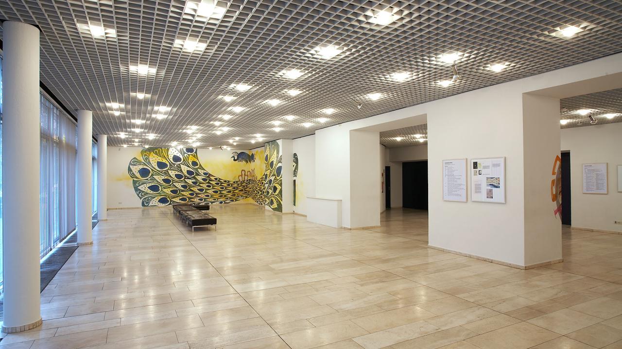 Spacious art gallery interior with a light tiled floor, white walls, and a colorful peacock mural. The ceiling has a grid design with evenly spaced lights.