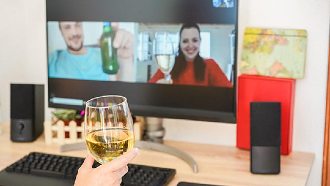 Eine Person hält ein Glas Weißwein vor einem Computerbildschirm, auf dem zwei Personen während eines Videogesprächs mit Getränken anstoßen.
