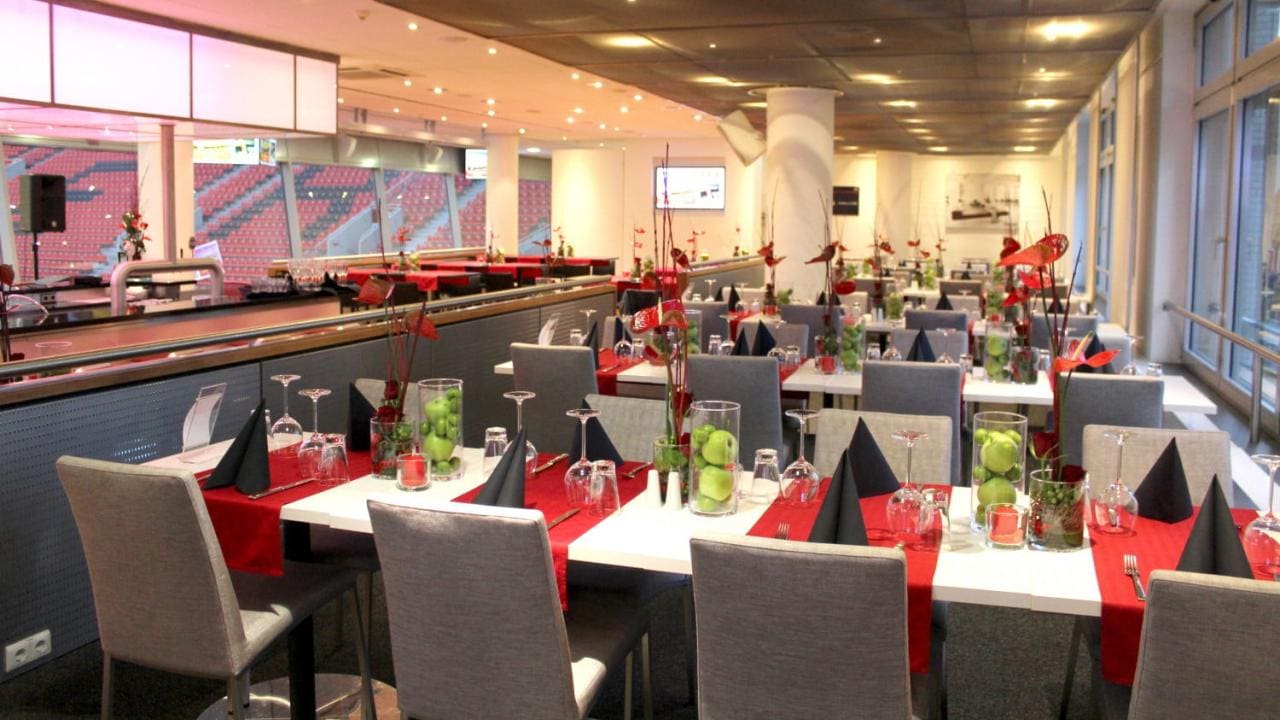 A modern restaurant interior with tables set for dining, featuring red and black accents, neatly folded napkins, and decorative centerpieces in glass vases. Large windows line the right side.