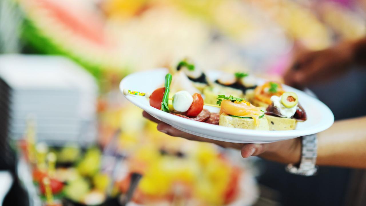 A hand holding a white plate with assorted appetizers, including tomatoes, cheese, salmon, and herbs.