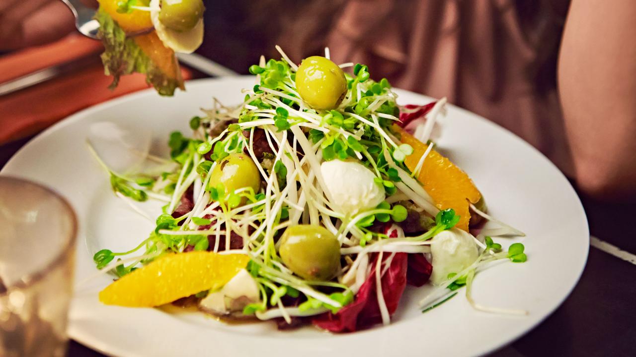 A salad with greens, olives, orange slices, sprouts, and mozzarella balls on a white plate. A fork with a bite of salad is being held above the plate.
