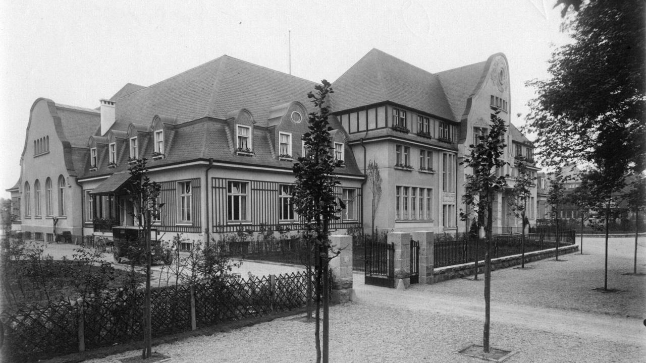 Historic building with multiple gabled roofs, surrounded by a fenced courtyard and young trees, set on a gravel path.