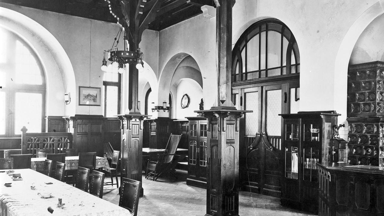 A vintage dining room with ornate wooden furniture, a long table with chairs, high ceilings, and arched windows and doorways.
