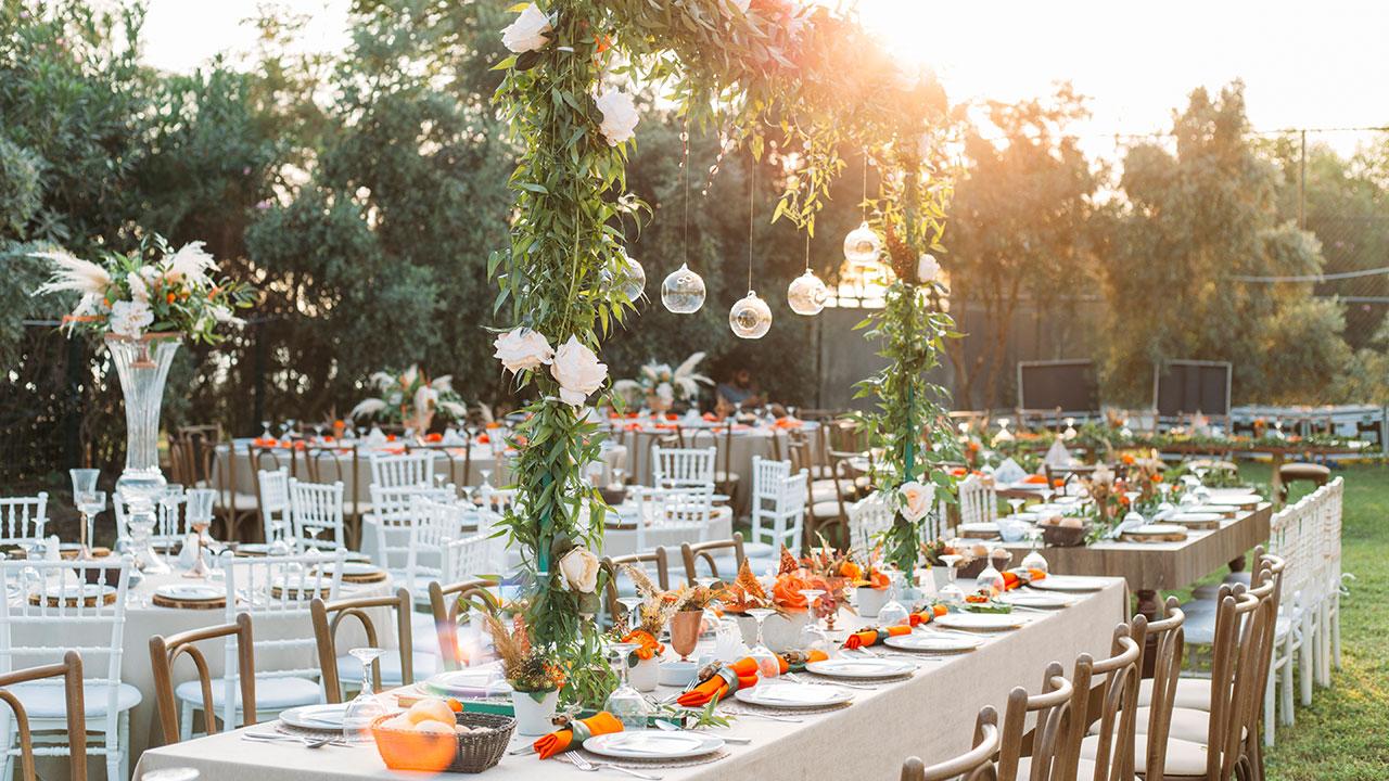 Outdoor wedding reception setup with long tables, white tablecloths, and floral centerpieces. Greenery and hanging glass ornaments adorn the area. Sunlight filters through surrounding trees.