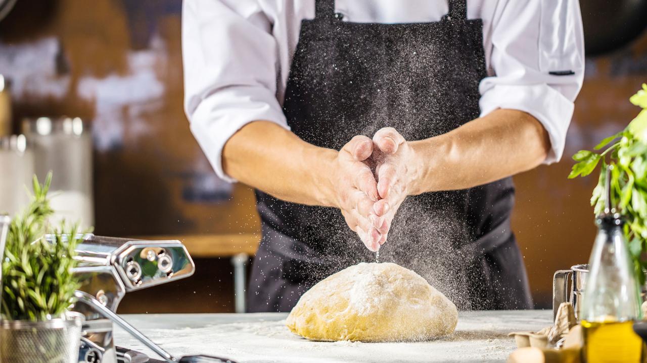 Ein Koch in weißem Hemd und schwarzer Schürze streut Mehl auf einen Teigballen auf einer Küchentheke, umgeben von Kräutern und Kochzutaten.