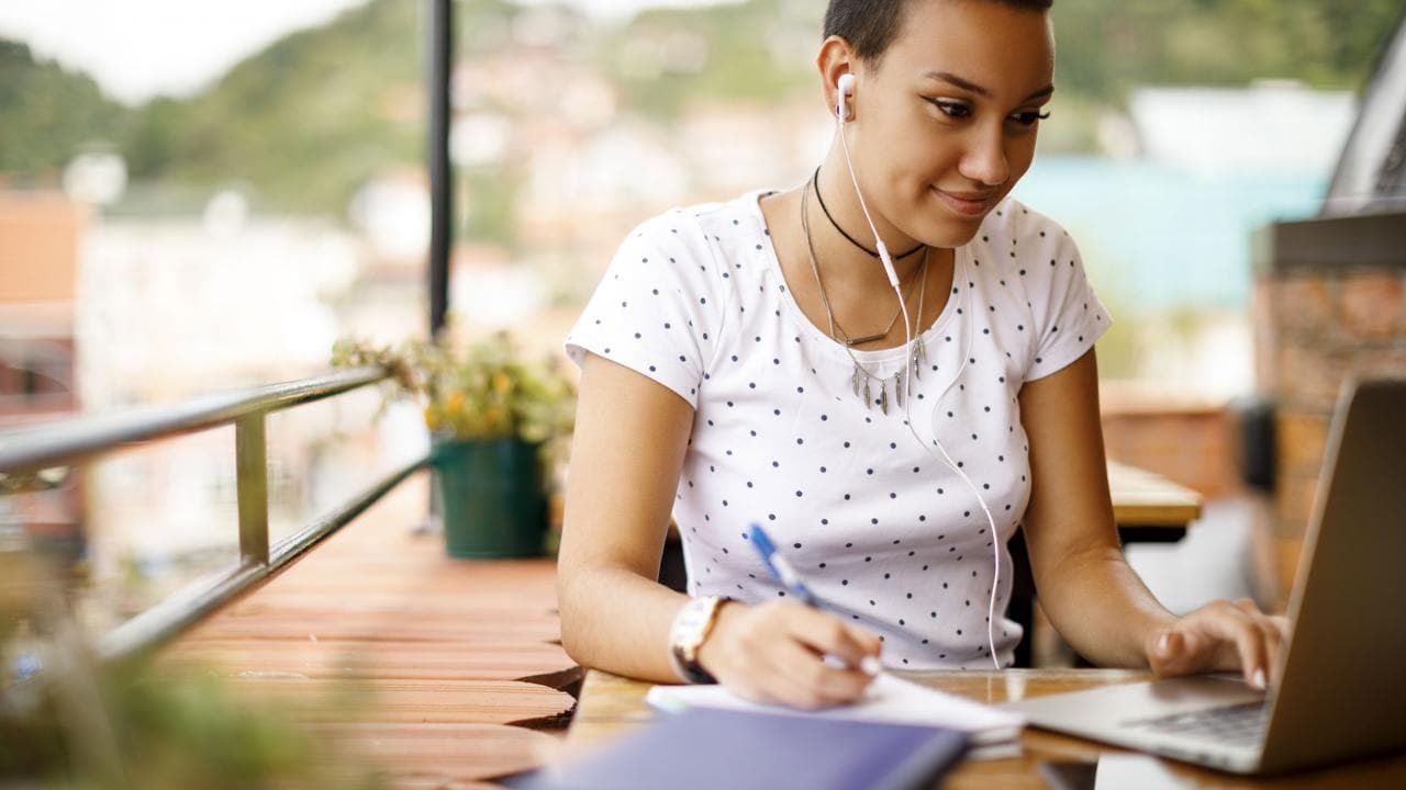 Person with short hair wearing earphones, working on a laptop and taking notes, sitting outdoors at a wooden table.