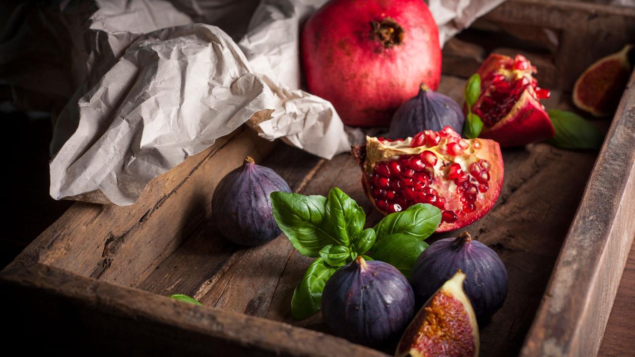 A wooden tray holds a whole pomegranate, a cut pomegranate, fresh figs, and basil leaves on crumpled paper.