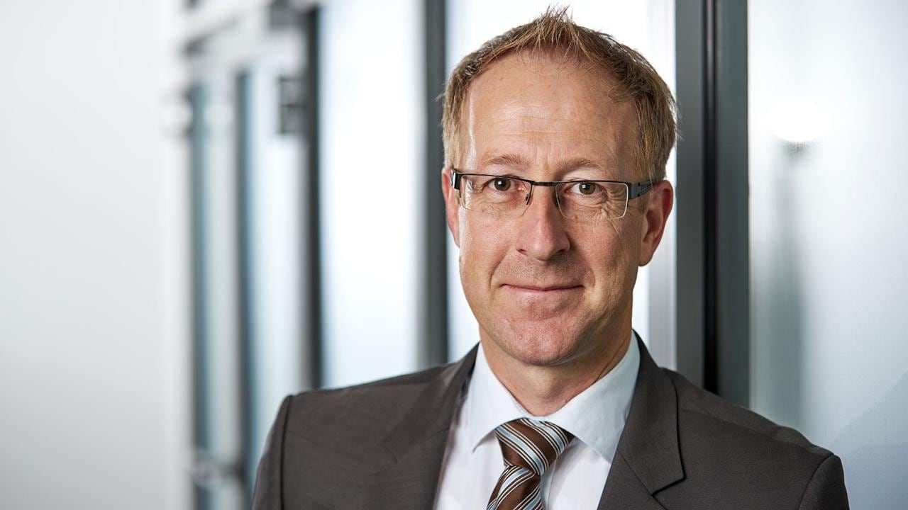 A man in glasses and a suit stands indoors, leaning against a glass wall.