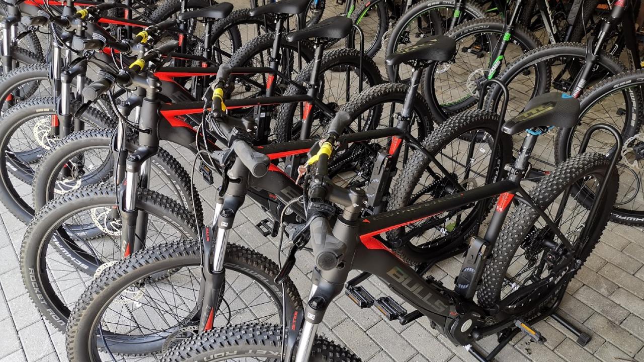 A row of mountain bikes with black and red frames are lined up on a brick pavement. More bikes are visible in the background.