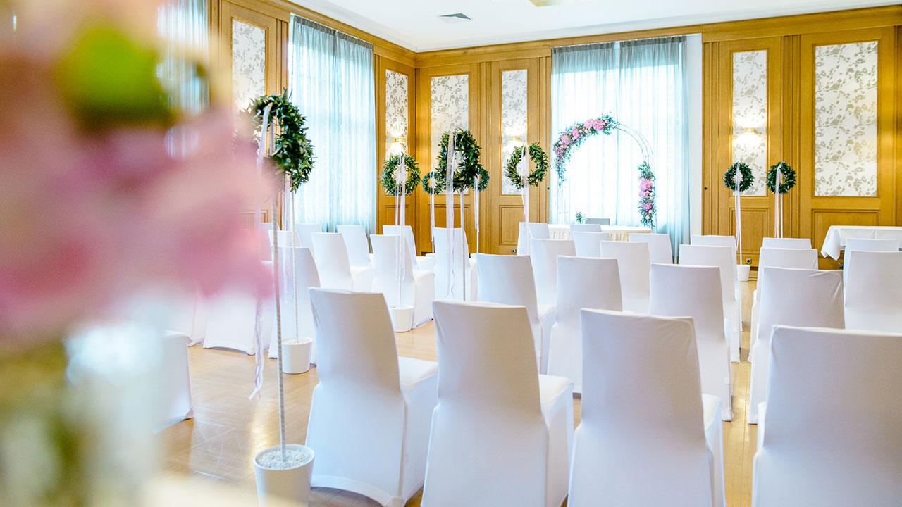 A brightly lit wedding ceremony room with rows of white chairs facing a decorated archway adorned with floral arrangements and greenery.