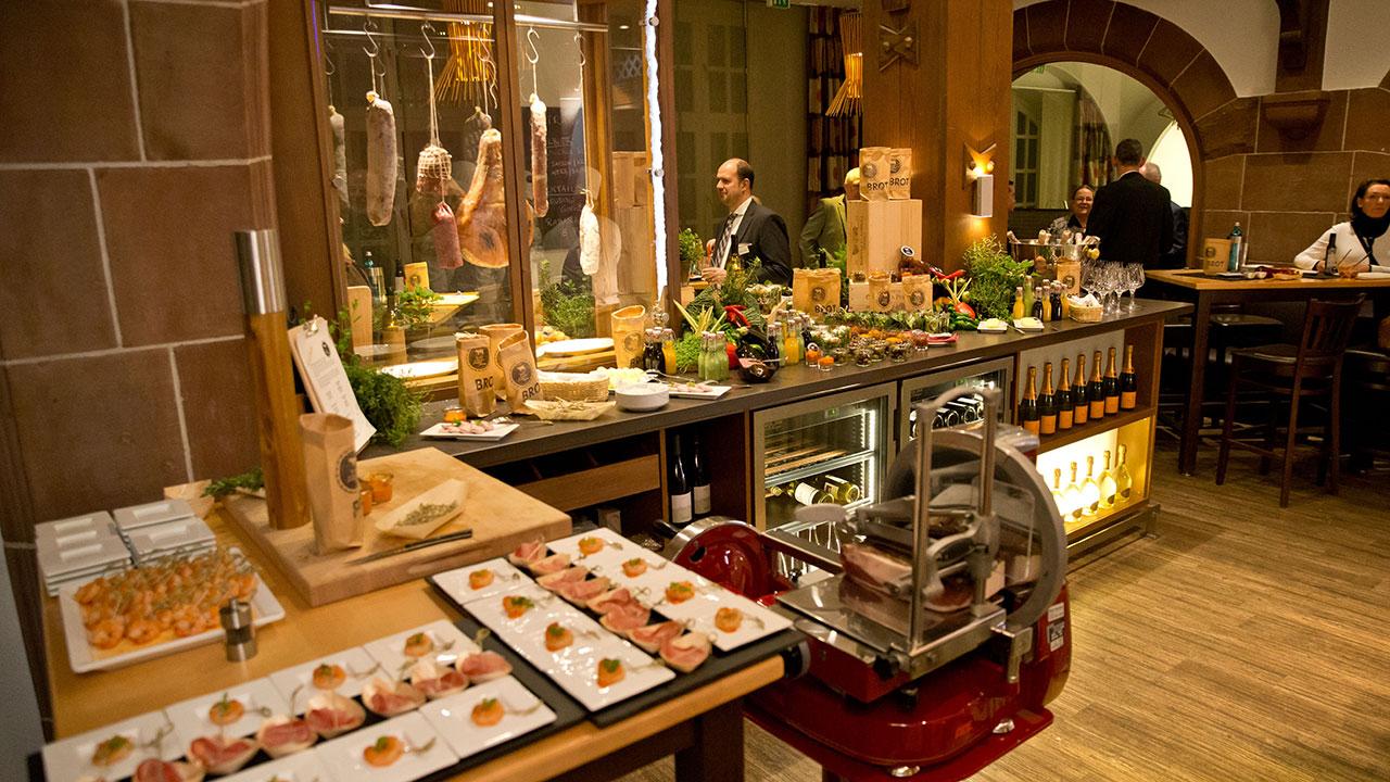 A deli-themed restaurant interior featuring cured meats hanging, a meat slicer, wine bottles on display, and appetizers arranged on plates, with customers seated at tables.