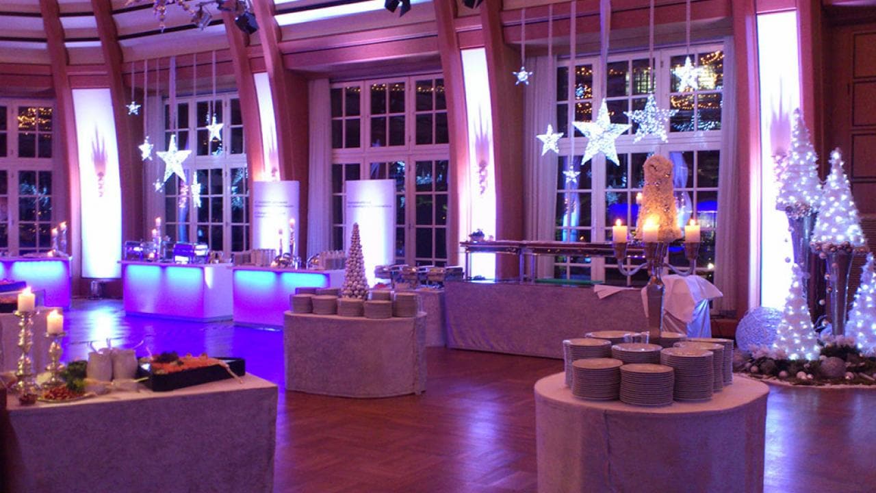A decorated event hall with round tables, stacked plates, and purple lighting. Star-shaped lights and festive decorations adorn the space.