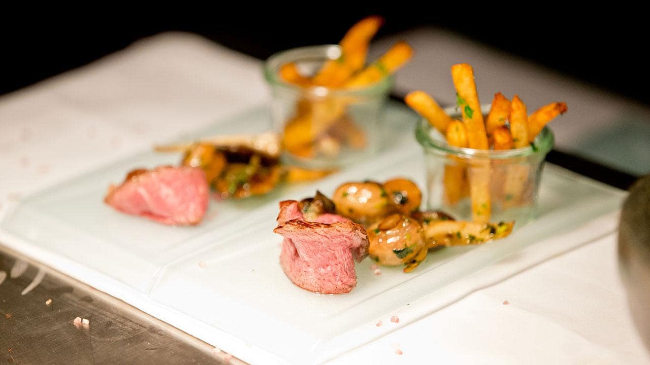 Two servings of sliced beef with sautéed mushrooms and herbs alongside glass jars of thin fries on a white serving tray.