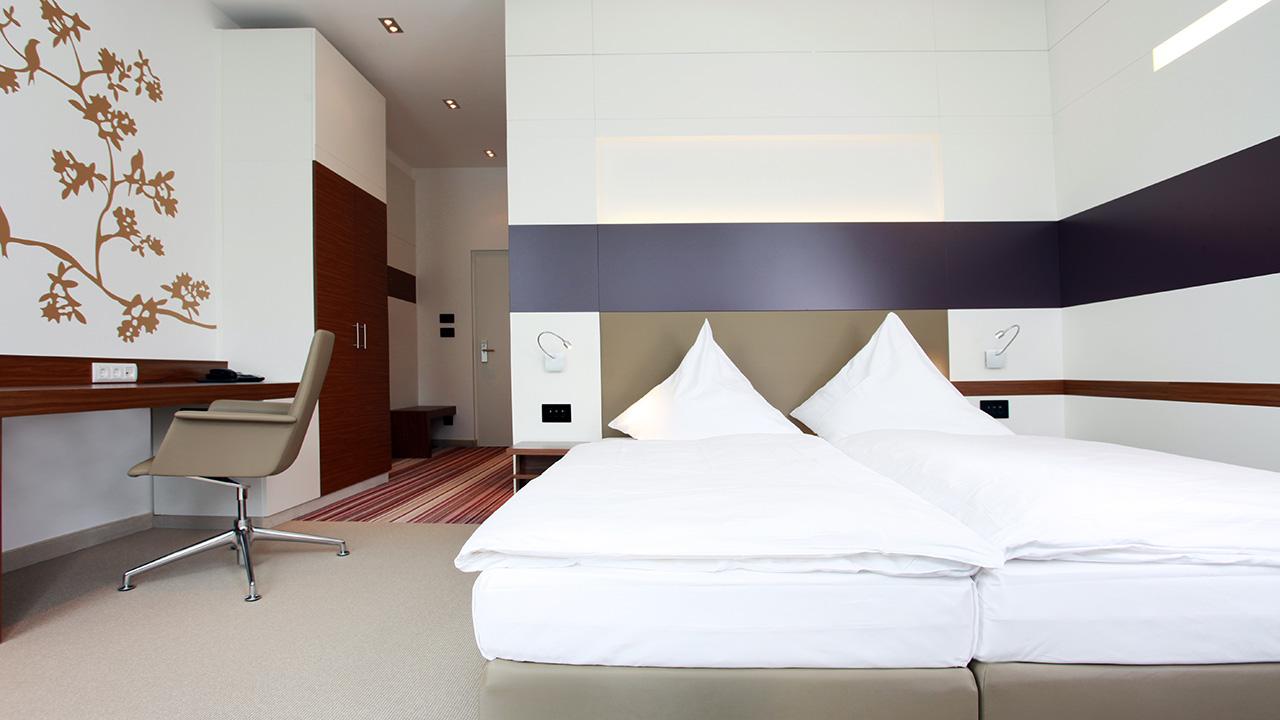 A modern hotel room with a large bed, a stylish chair at a desk, and contemporary wall decor featuring a tree design. The walls are predominantly white with brown and purple accents.