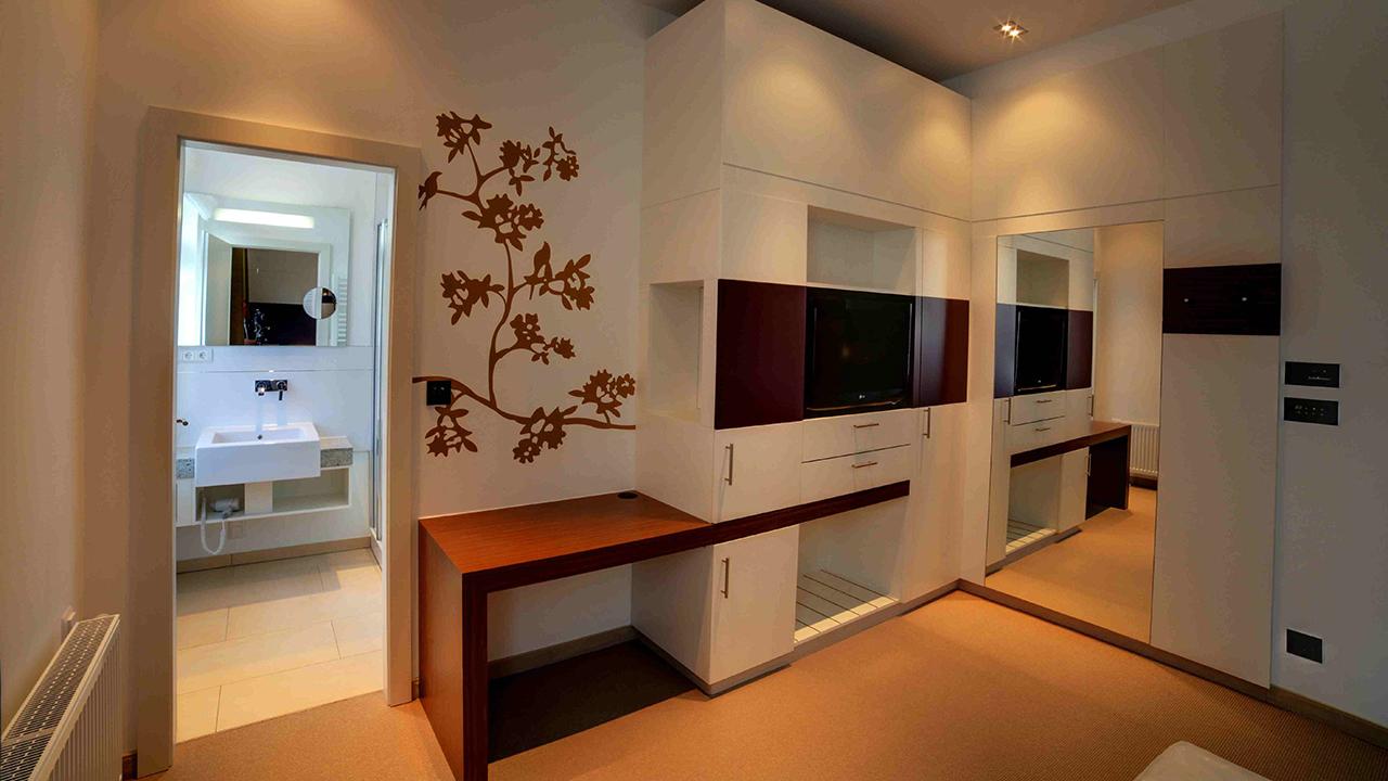 Modern room with white cabinetry, a wall-mounted TV, and a wooden desk extending from storage. Decorative floral wall art is present. An open door leads to a bathroom with a sink and mirror.