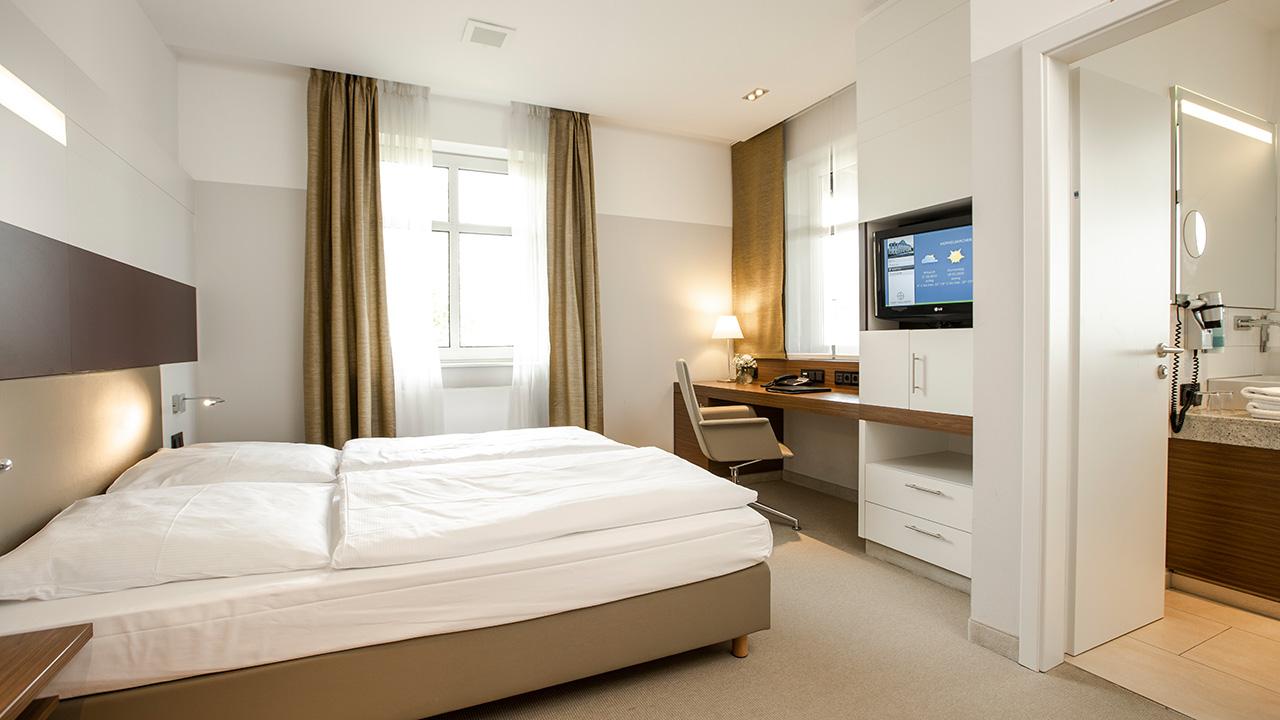 A modern hotel room with a double bed, desk, wall-mounted TV, and a glimpse of a bathroom. Beige and white decor with natural light from a window.