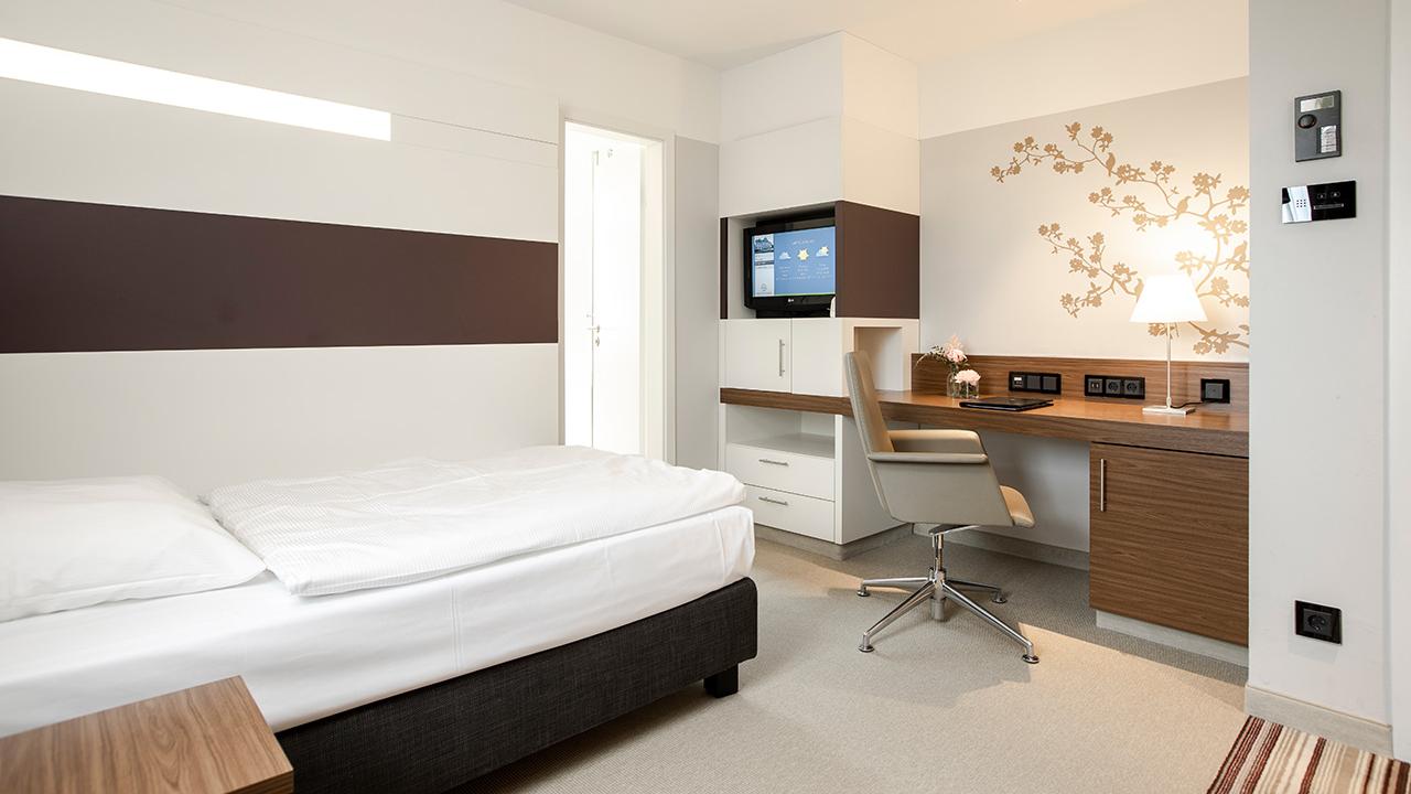 Modern hotel room with a single bed, desk, swivel chair, wall-mounted TV, and floral wall art. Doorway to a brightly lit area is visible.