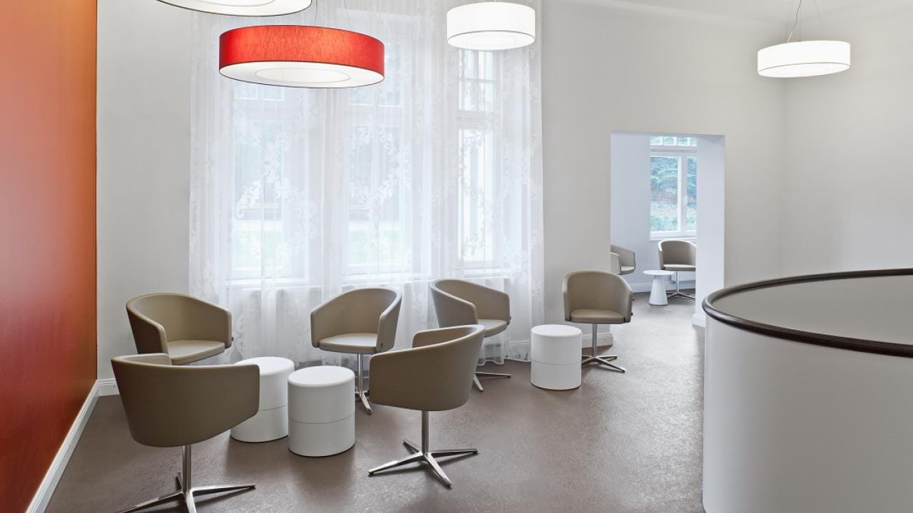Bright waiting area with modern chairs, small round tables, and large windows with sheer curtains. Ceiling lights provide ample illumination.