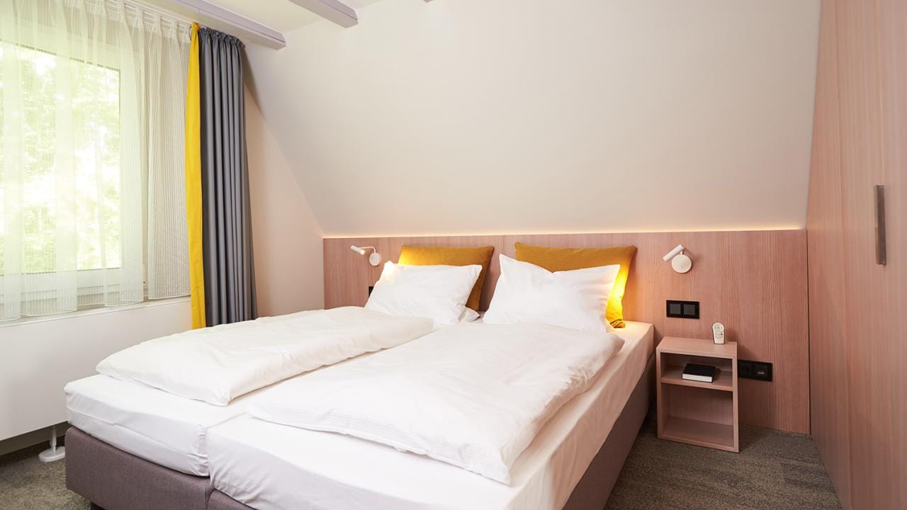 A modern bedroom with two single beds pushed together, white bedding, wooden nightstand, wall-mounted lamps, and a window with sheer and gray curtains.