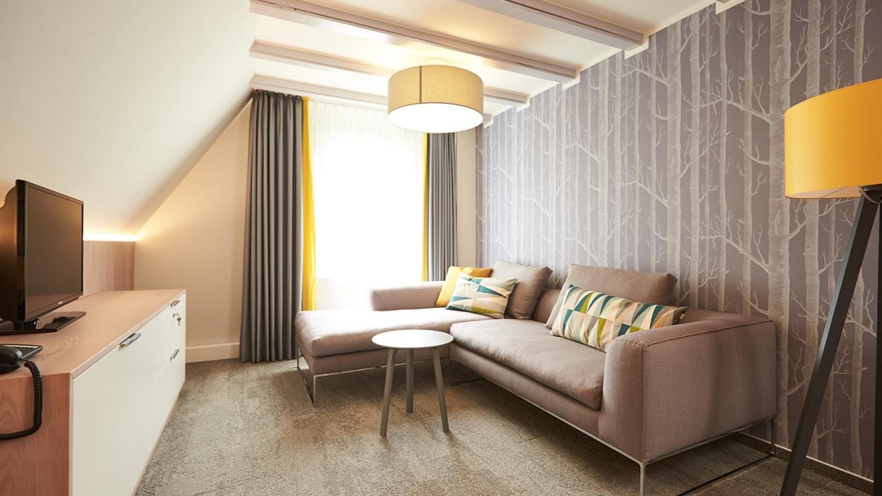A modern living room with a gray L-shaped sofa, patterned pillows, a round table, a TV, and a floor lamp. The walls have tree-patterned wallpaper, and a window with curtains is visible.