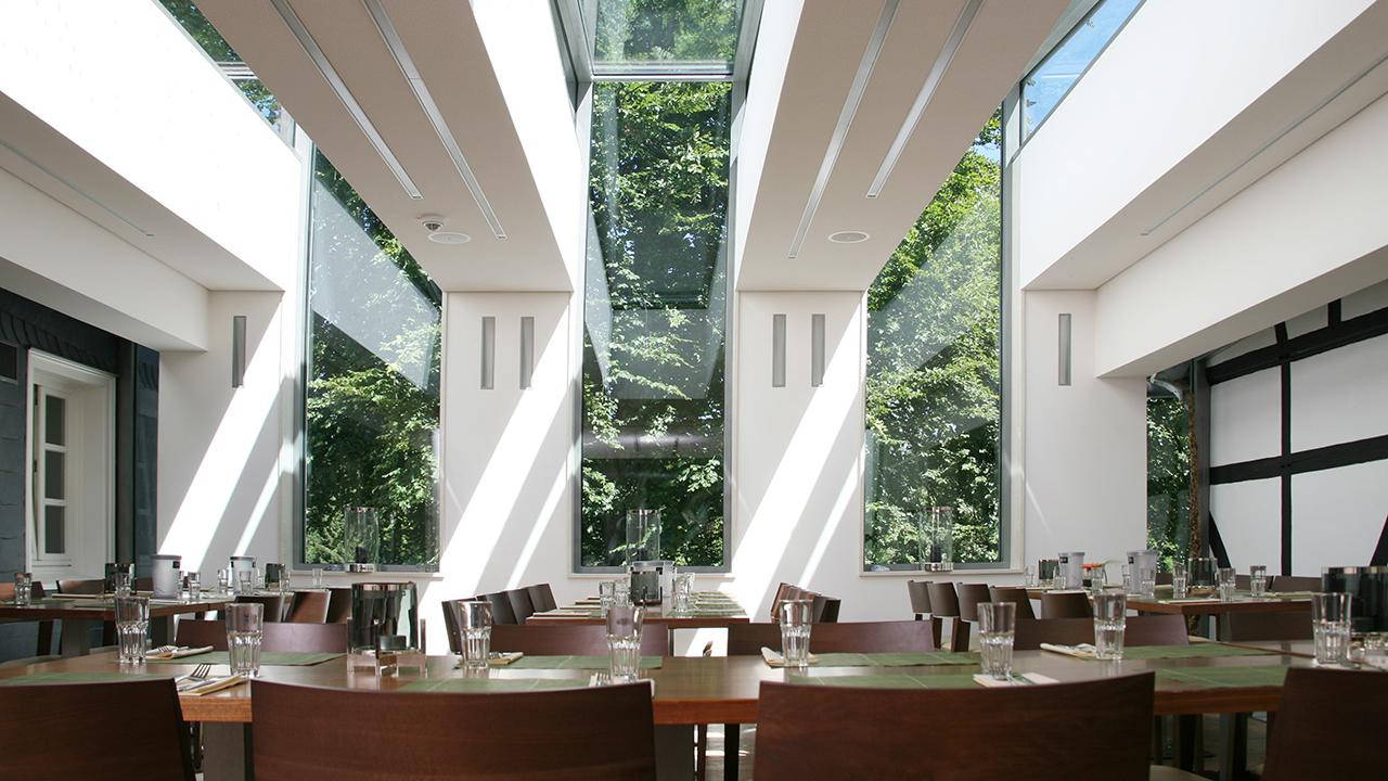 A modern restaurant interior with large windows and skylights, featuring wooden tables, neatly arranged chairs, and table settings with glasses and placemats. Greenery is visible outside.