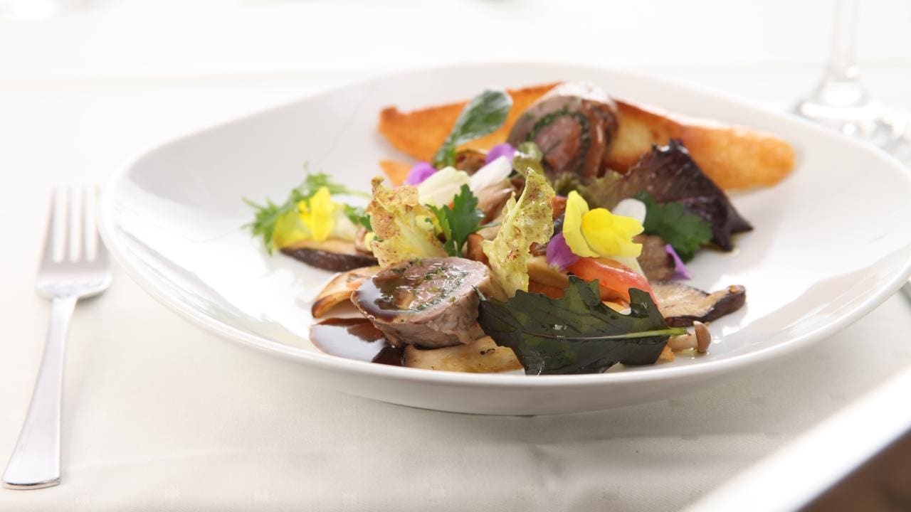 A gourmet dish with meat, edible flowers, and greens arranged on a white plate, accompanied by a fork on a tablecloth.