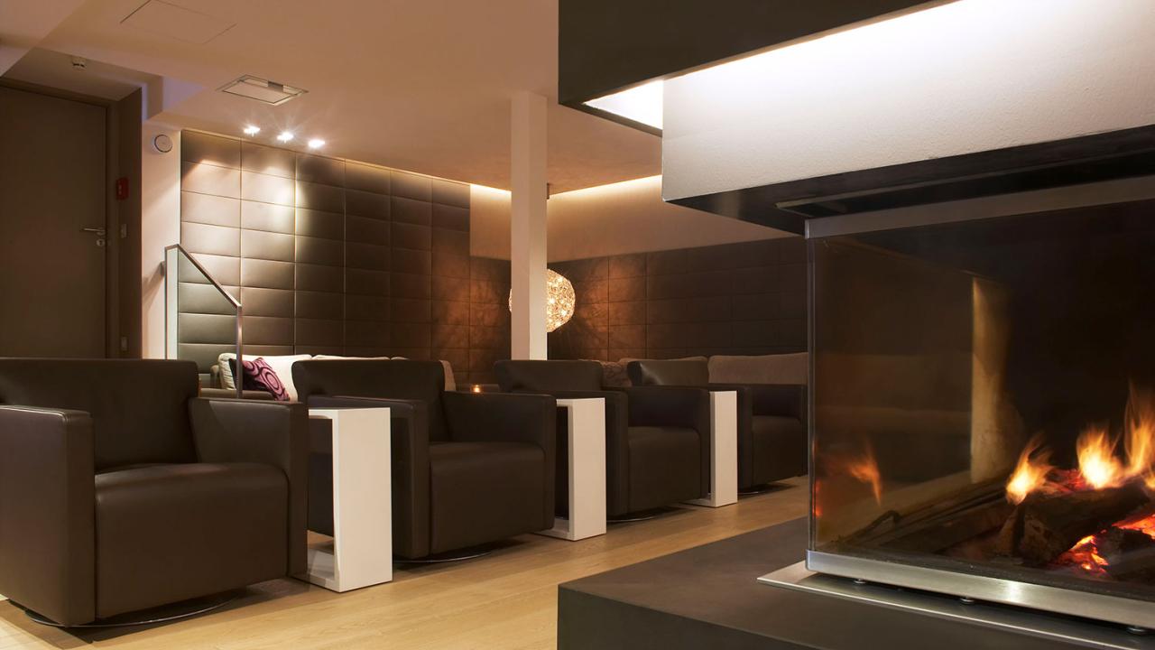 A modern lounge area with brown armchairs and white side tables facing a glass-enclosed fireplace. The room has dim lighting and a sleek, minimalist design.