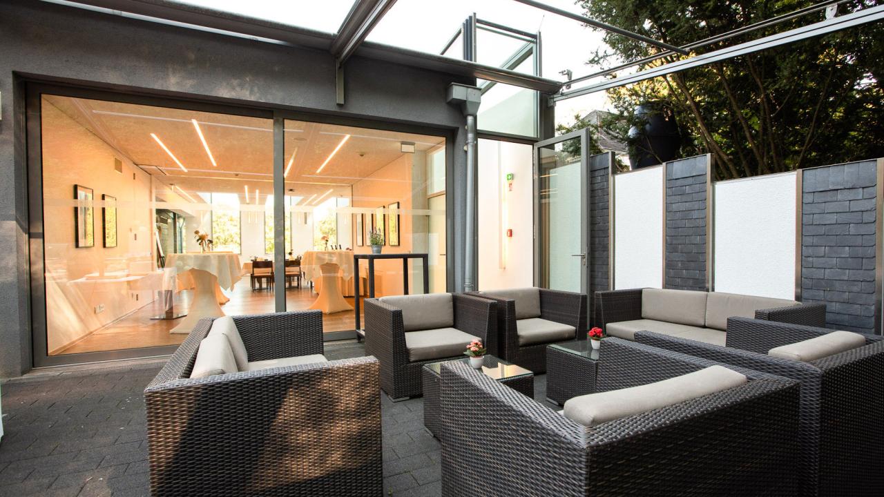 Outdoor seating area with wicker sofas and tables, adjacent to a modern glass-walled room set for dining.