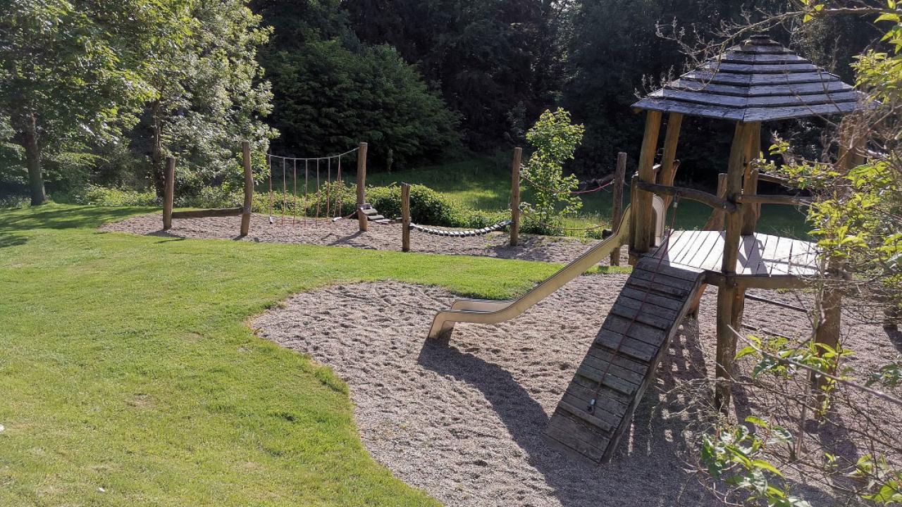 Playground with a wooden slide and climbing structure on a grassy area, surrounded by trees and bushes under a sunny sky.