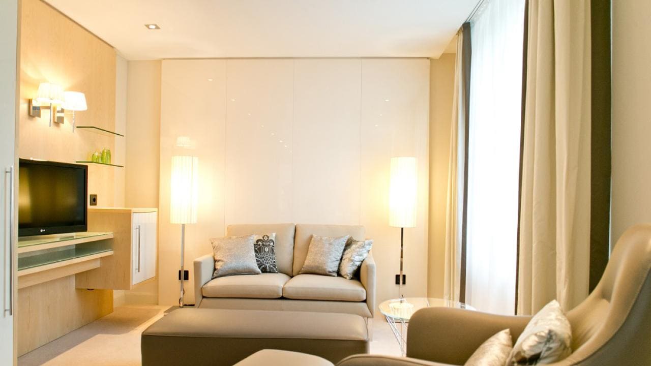 Modern living room with beige sofa, cushioned chair, ottoman, and mounted TV. Floor lamps and curtains complement the minimalist decor.