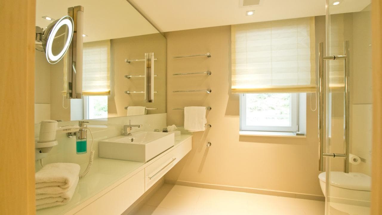 Modern bathroom with a large mirror, white sink, wall-mounted towel racks, and a window with a blind. Bright lighting and neutral tones create a clean and spacious feel.