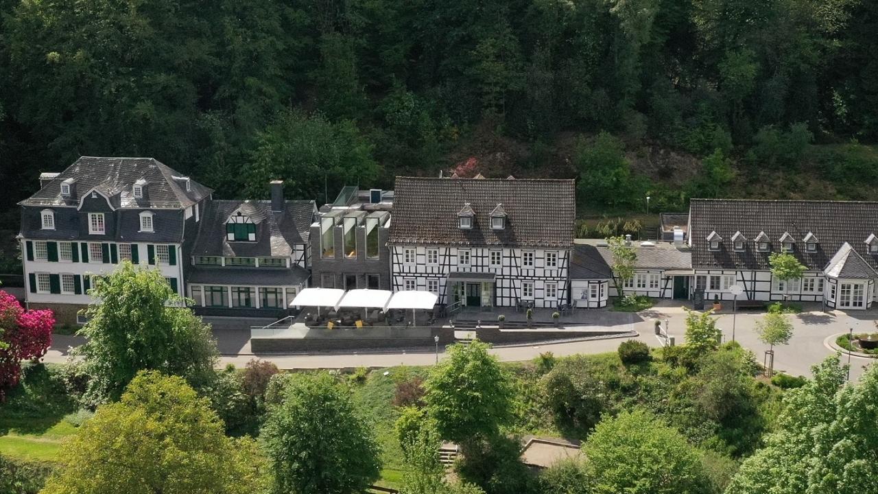 Luftaufnahme eines historischen Gebäudekomplexes, umgeben von üppigem Grün und Bäumen. Das Gebäude weist eine klassische Architektur mit einer Mischung aus Backstein- und Holzelementen auf.