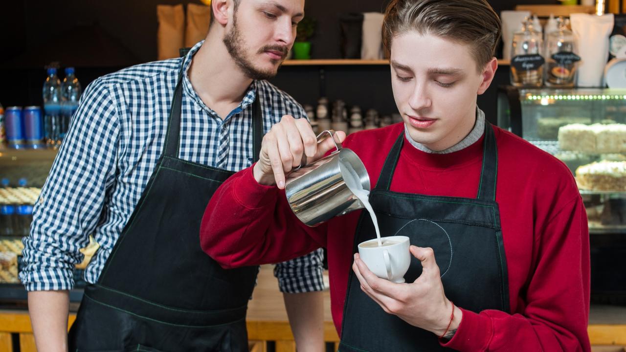 Two baristas in a cafe. One pours milk into a cup, while the other looks on. They wear black aprons and stand in front of a counter with pastries and a coffee machine.