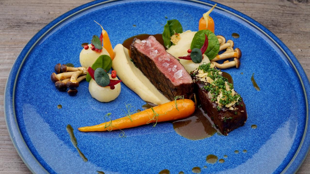 A plated gourmet dish featuring beef, mashed potatoes, carrots, mushrooms, and sauce on a blue plate.