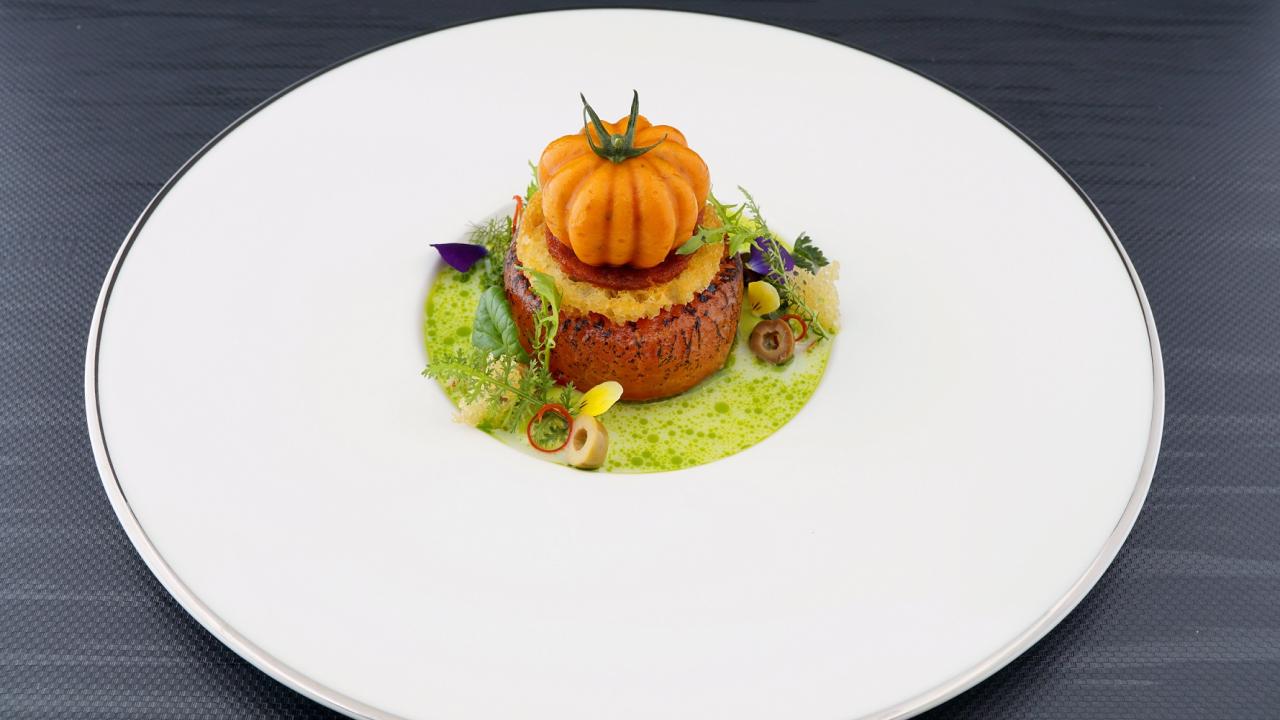 A gourmet dish featuring a small pumpkin on a round patty, garnished with herbs, flowers, and a green sauce, served on a white plate with a dark background.
