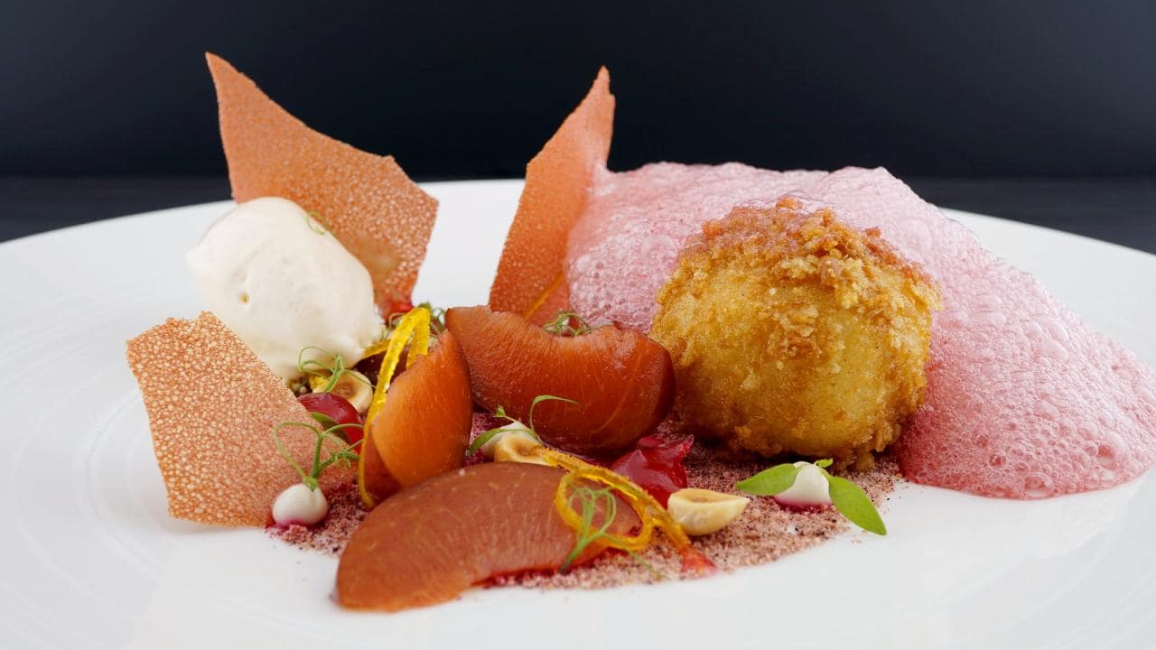A plated dessert with poached pears, a fried ball, pink foam, white ice cream, and decorative crispy shards on a white plate.