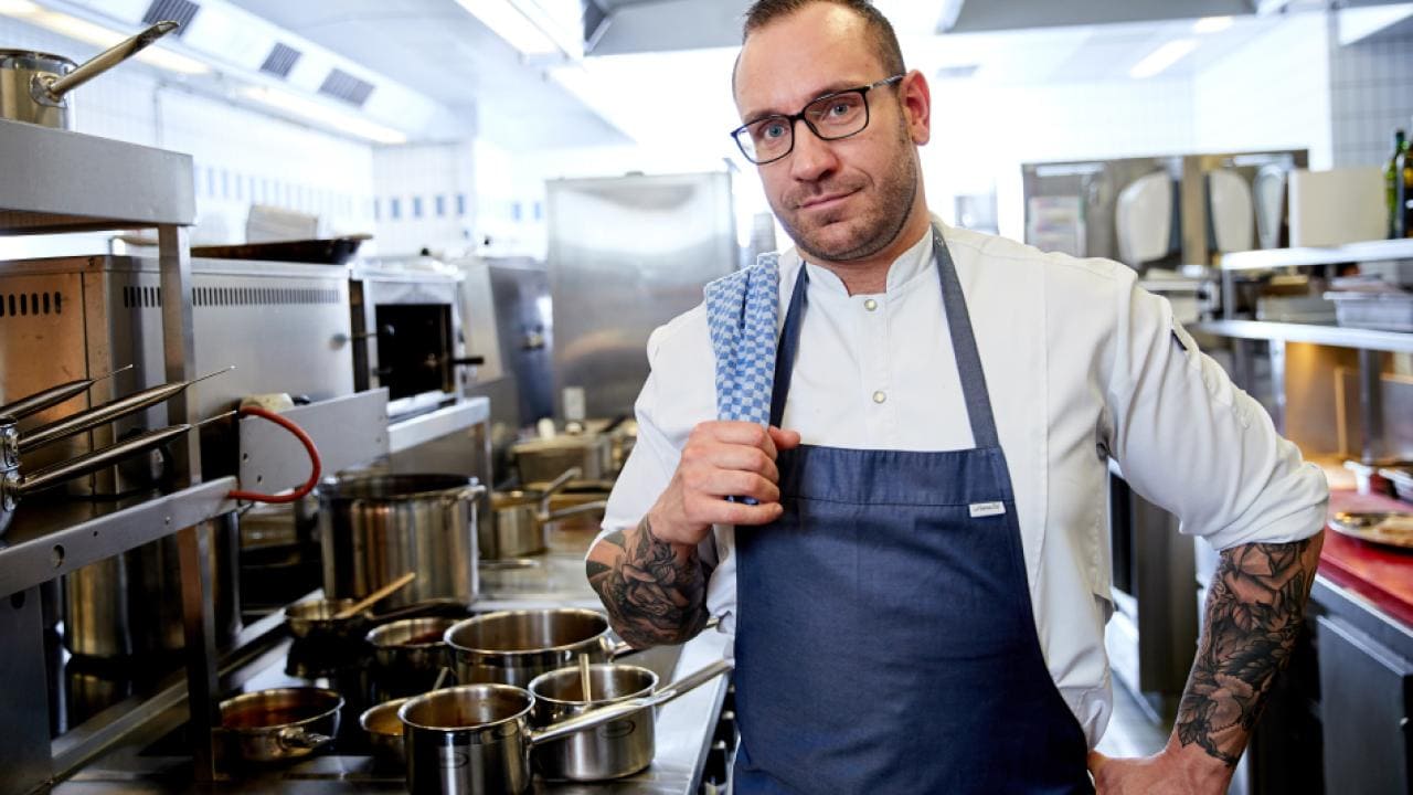 A chef in a white coat and blue apron stands confidently in a professional kitchen, with various pots and cooking utensils around him.