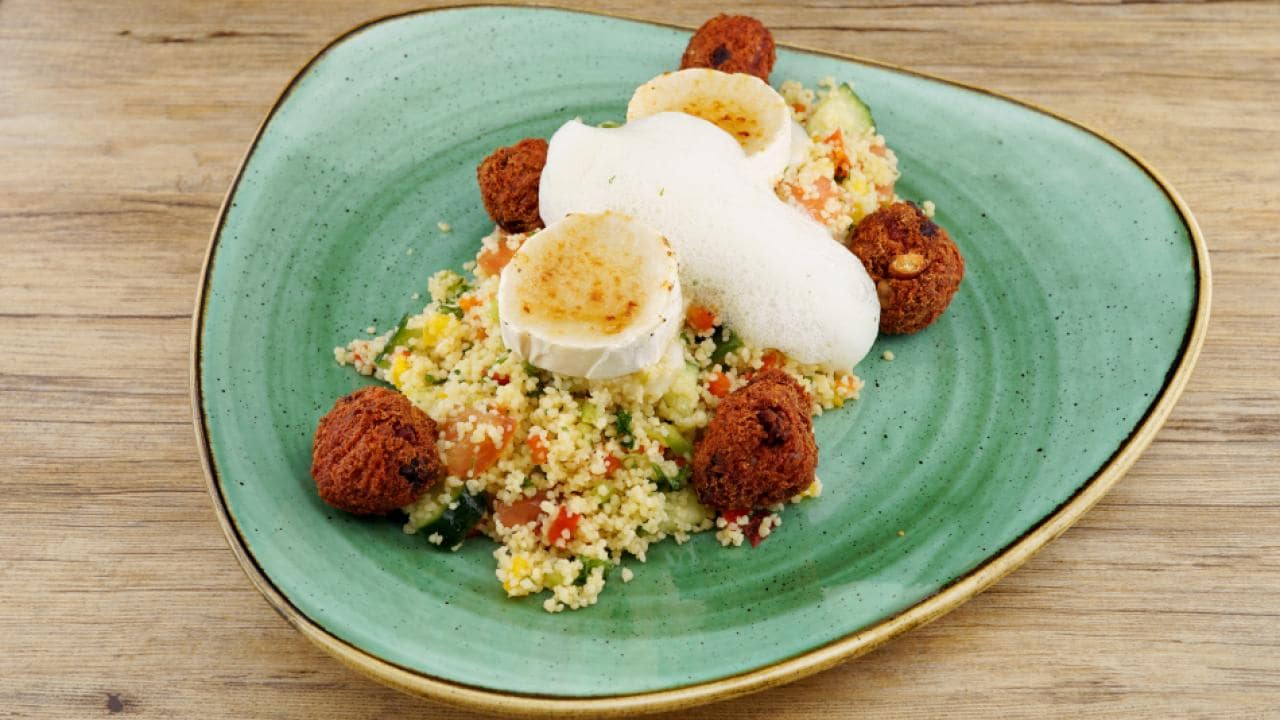 A green plate with couscous salad topped with falafel balls, goat cheese slices, and a white foam garnish.
