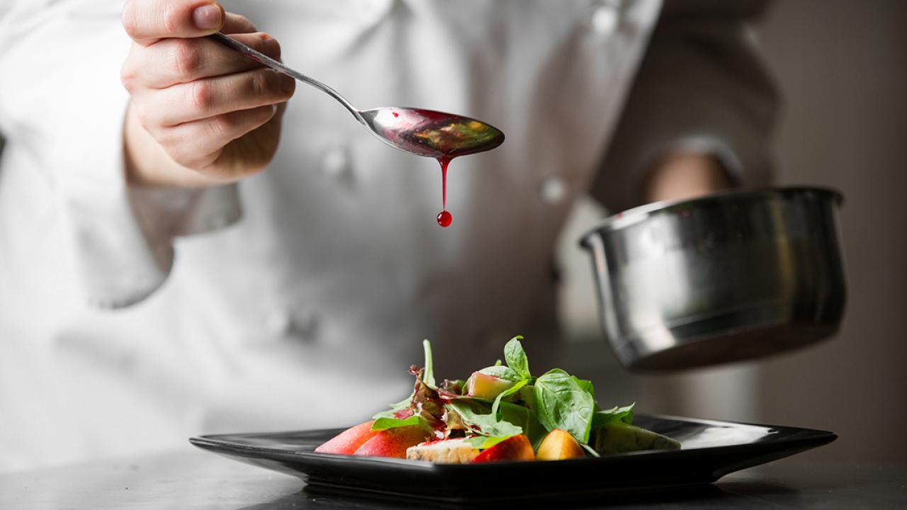 A chef in a white coat drizzles sauce from a spoon onto a dish of mixed greens and fruit on a black square plate.