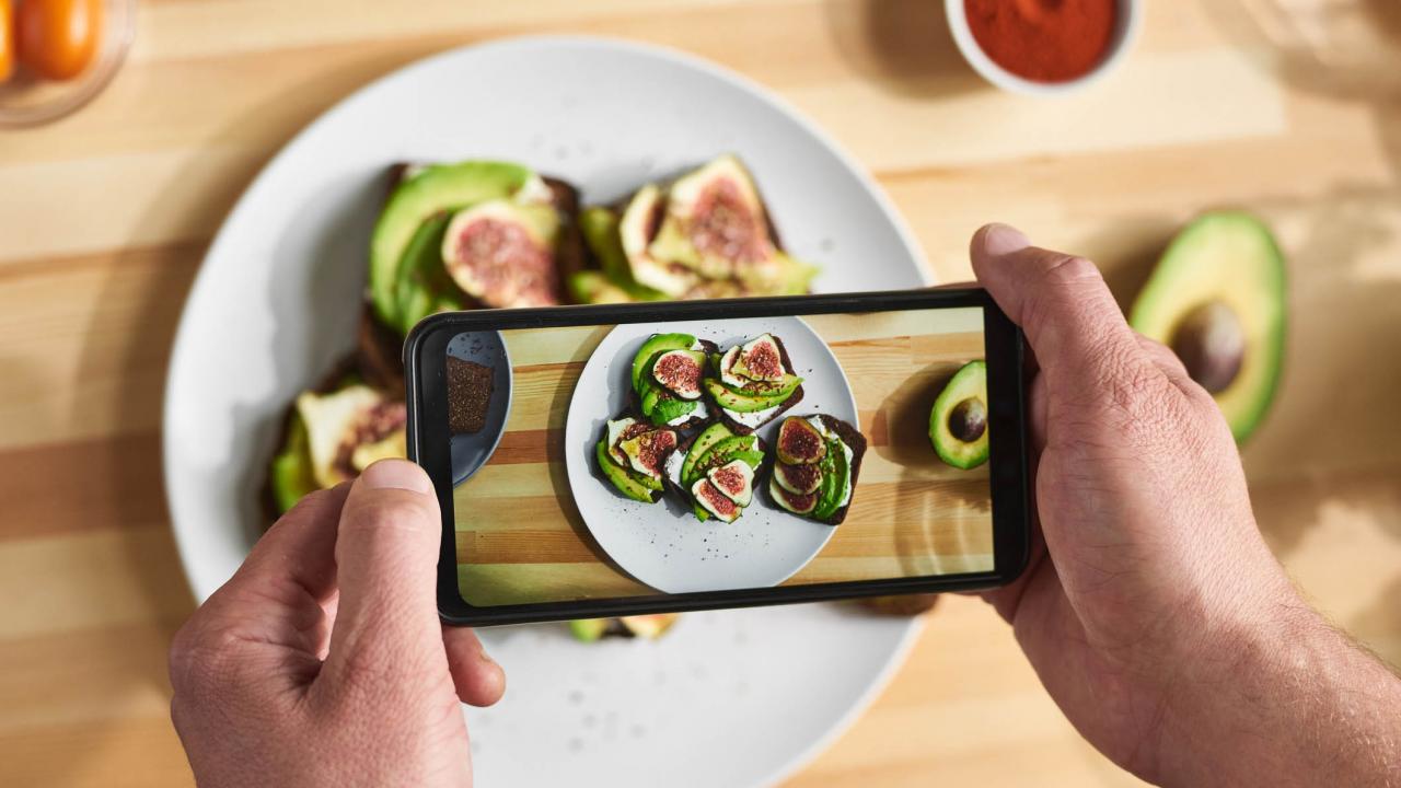 Person taking a photo of avocado and fig slices on toast with a smartphone, on a striped wooden table.